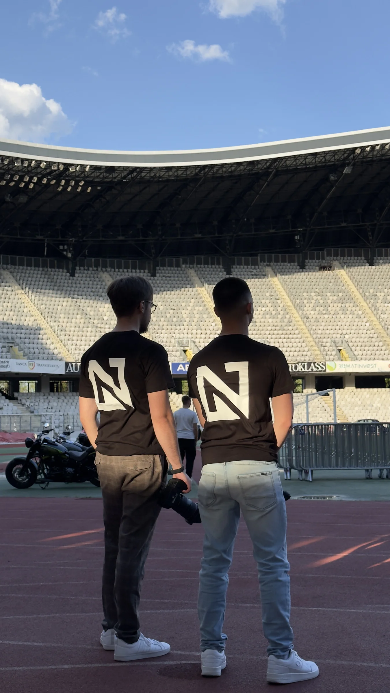Norris and Zoard wearing NZ shirts, seen from behind at a stadium, with a camera in hand.