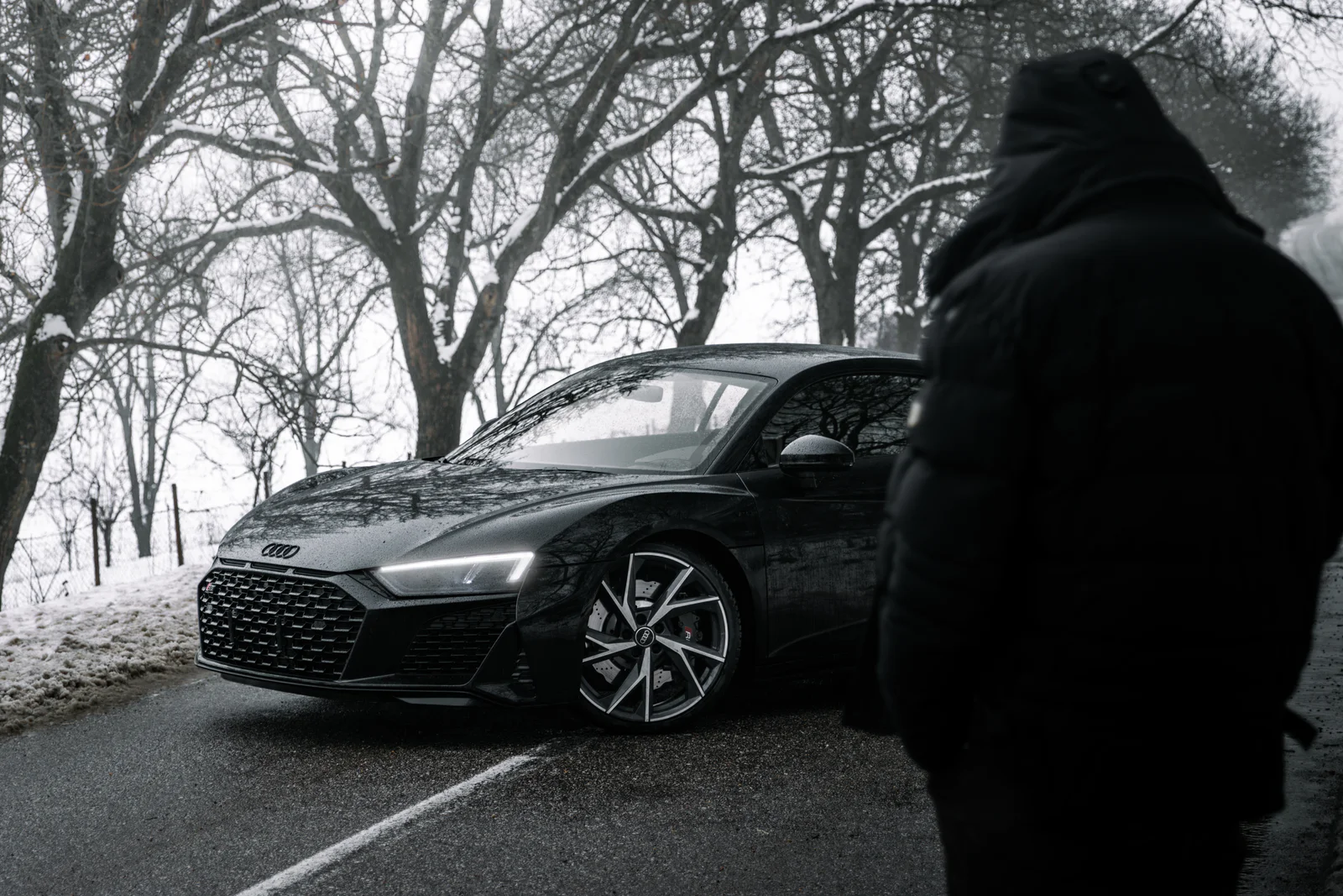 A hooded figure in the foreground beside the R8 on the winter road