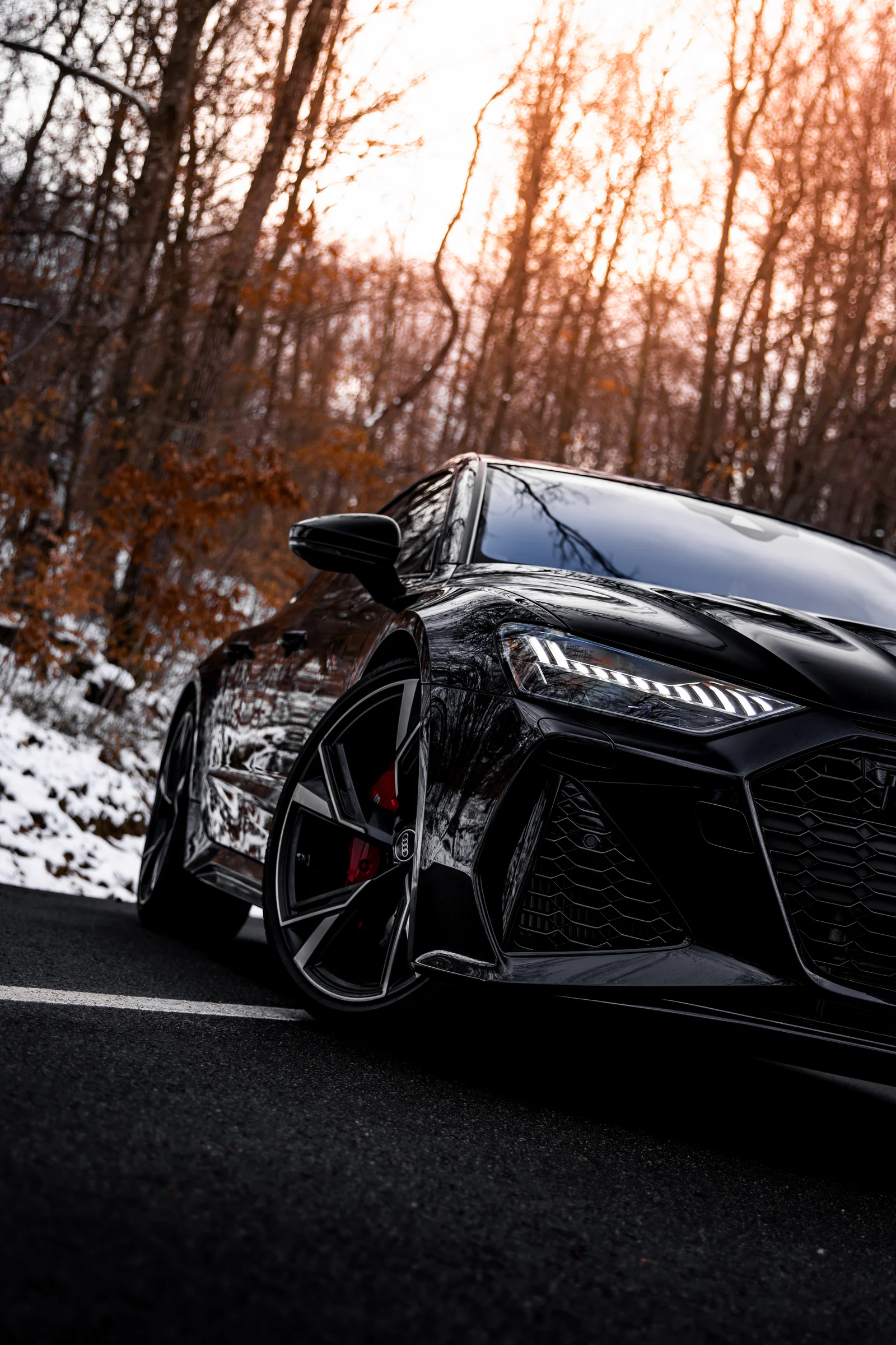Low-angle Audi RS7 headlight and front wheel beneath sunlit trees